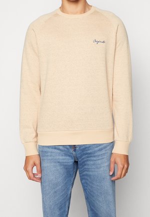 Pfirsichfarbener Pullover aus strukturiertem Stoff mit gerippten Bündchen und Saum, versehen mit einem kleinen blauen Logo auf der linken Brust, kombiniert mit blauen Jeans.