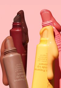 Drei Quetschbehälter mit Lippenpflege in Braun, Rot und Orange, jeweils mit einem glänzenden Finish und einer tropfenden Textur, vor einem rosa Hintergrund.