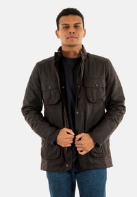 Bruine gewaxte katoenen jas met een ritssluiting en drukknopen, twee voorkruimelpockets en een klassieke kraag, gedragen over een zwart shirt.