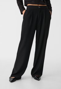 Zwarte wijde broek gedragen met zwarte puntige flats, handen in de zakken, en een zwarte cropped jas zichtbaar op taillehoogte.