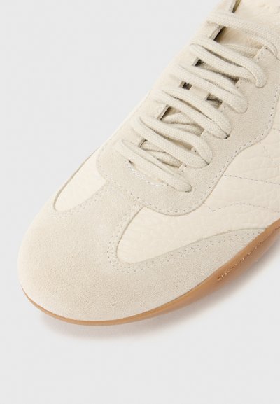 Μπεζ παπούτσι από σουέντ με υφασμένους κρεμ χρωματισμούς, στρογγυλή μύτη και κρεμ κορδόνια. Επίπεδη σόλα από καουτσούκ με διακριτικές ραφές.