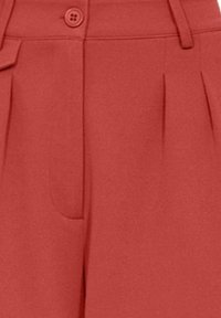 Rode, op maat gemaakte broek van soepel materiaal, met een voorzak, plooien en een knoopsluiting in de taille.