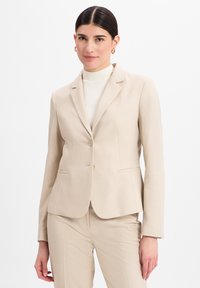 Blazer d'affaires beige sur mesure avec un motif texturé subtil, des revers en pointe, une fermeture à un bouton et des poches avant.