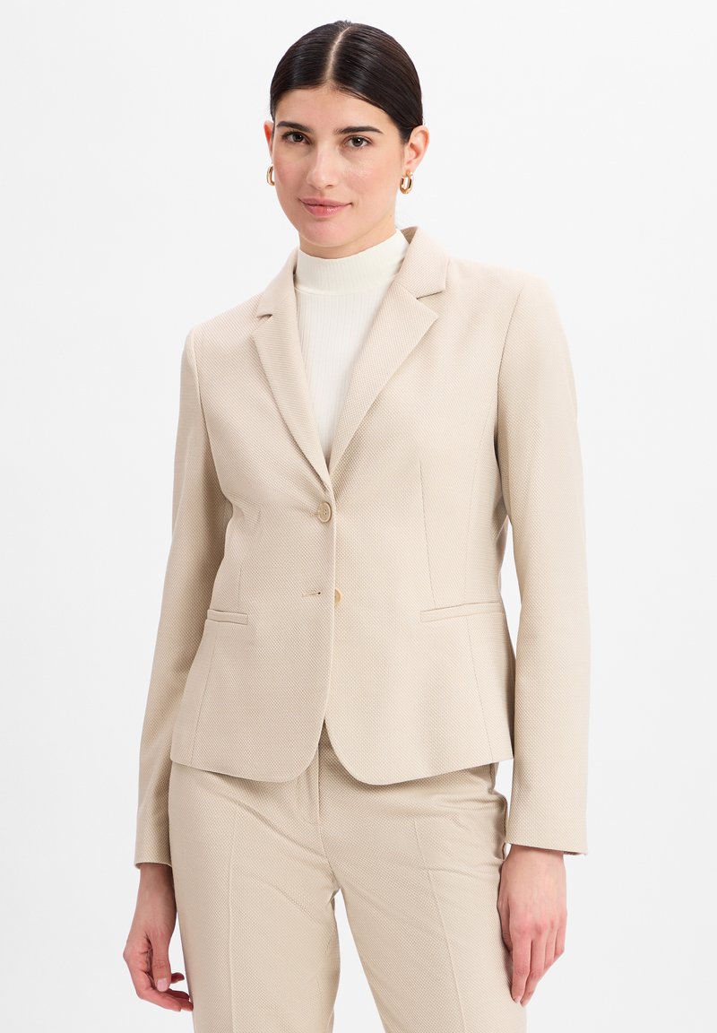 Blazer d'affaires beige sur mesure avec un motif texturé subtil, des revers en pointe, une fermeture à un bouton et des poches avant.