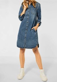 Kvinna som bär en knälång knäppt jeansklänning med uppvikta ärmar och vita högklackade sneakers, stående med en hand i fickan.