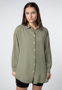 Olijfgroene overhemd met lange mouwen, knoopsluiting en een gestructureerde stof met een kreukelige uitstraling. Het heeft een puntig shirtkraag en zijsplitten.