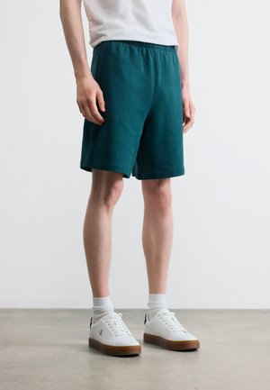 Bărbat purtând pantaloni scurți cu talie elastică verde închis, șosete albe și adidași albi cu tălpi maro, stând pe un podea netedă gri.