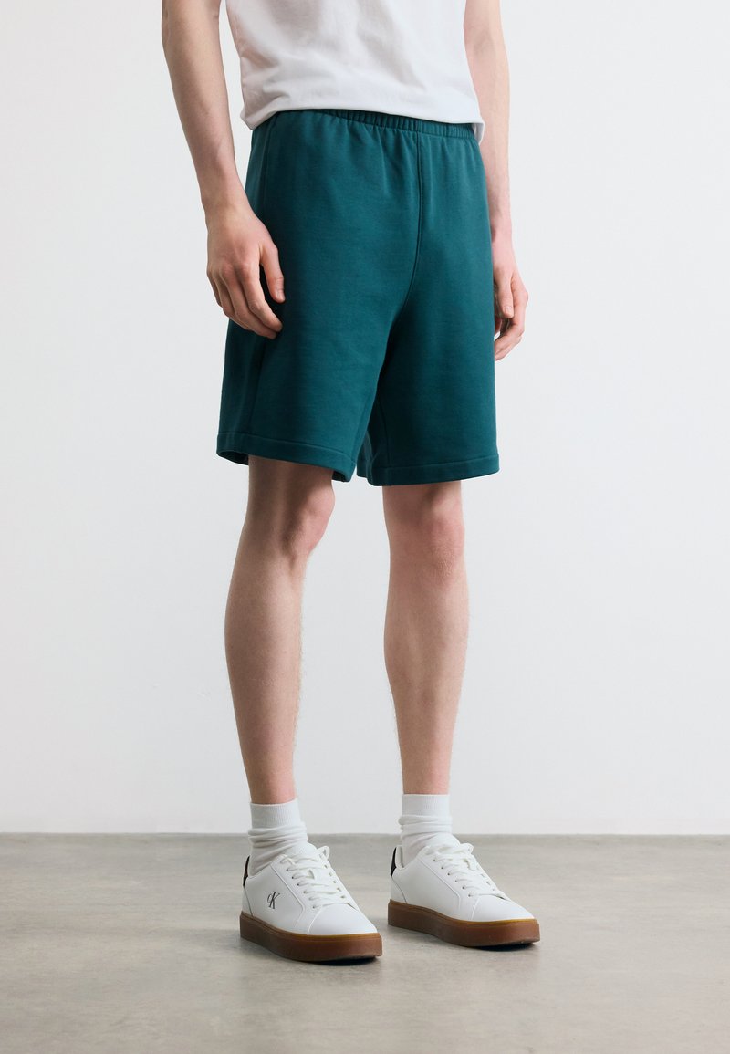 Άνδρας που φοράει σκούρα πράσινα σορτς με ελαστική μέση, λευκές κάλτσες και λευκά αθλητικά παπούτσια με καφέ σόλες, στέκεται σε λείο γκρι πάτωμα.