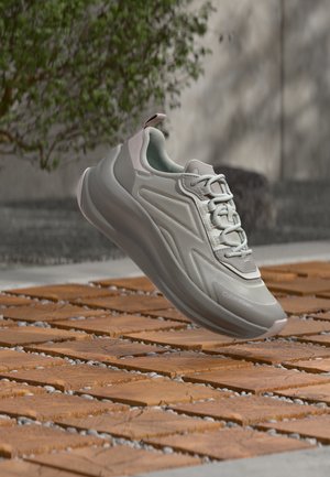 Lichtgrijze en witte sneaker met dikke zool en veters die boven een houten betegeld oppervlak buitenshuis zweeft, nabij een wazige boom en muur.