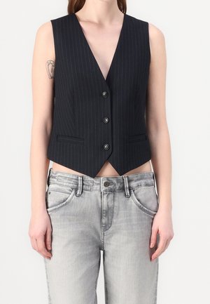 Osoba ubrana w czarną prążkowaną kamizelkę na guziki i jasnoszare, wyblakłe dżinsy, prezentująca tatuaż na górnej lewej ręce.