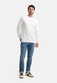 Hvit bomullssweatshirt med rund hals, ribbestrikkede mansjetter og kant; parret med lyseblå denimjeans og beige sneakers med svarte detaljer.
