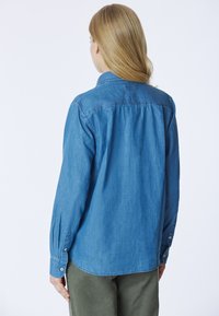 Camicia in denim azzurro chiaro con collo, maniche lunghe e polsini con bottoni. Presenta un orlo dritto e un dettaglio arricciato sul retro.