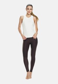 Giovane donna con una lunga coda di cavallo che indossa un top bianco senza maniche e leggings scuri, in piedi a piedi nudi contro uno sfondo chiaro e semplice.