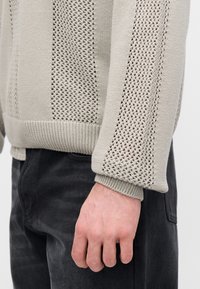 Μπεζ πουλόβερ πλεκτό με λεπτομέρειες ανοιχτής πλέξης στα μανίκια και στο σώμα, με ριμπ μανσέτες και ρεβέρ. Φορεμένο πάνω από σκούρο γκρι παντελόνι.