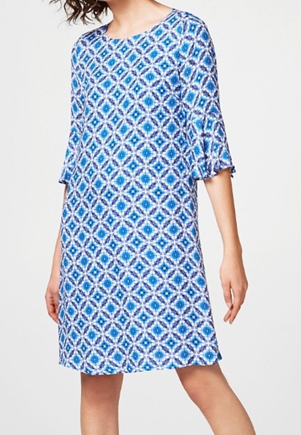 Femme portant une robe mi-longue bleu et blanc à motifs géométriques avec des manches à volants aux coudes sur fond uni.
