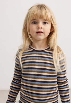 Jeune enfant aux longs cheveux blonds portant un t-shirt à manches longues rayé bleu, marron, beige et noir, debout devant un fond clair uni.
