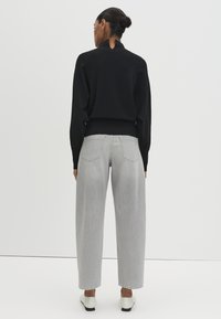 Maglione nero con collo alto e dettaglio cut-out, abbinato a jeans a gamba larga grigio chiaro. Il modello indossa scarpe bianche senza lacci.