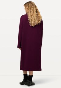 Langer lila Mantel mit strukturiertem Muster, weiten Ärmeln und gerader Silhouette, kombiniert mit schwarzen Knöchelstiefeln.