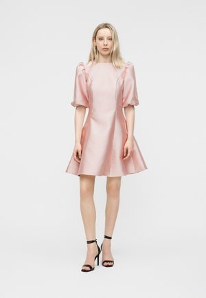 Mujer de pie con un vestido de raso rosa pálido con mangas abullonadas y sandalias negras de tacón alto, sobre un fondo blanco liso.