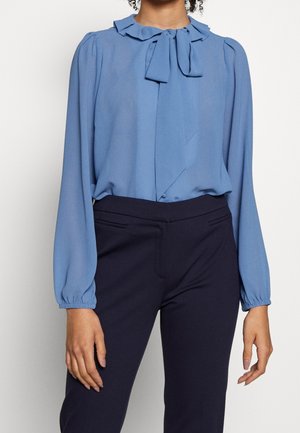 Kobieta ubrana w jasnoniebieską bluzkę z długim rękawem, falbankowanym kołnierzykiem i kokardą oraz ciemnogranatowe dopasowane spodnie na jednolitym tle.