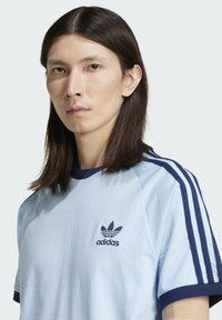 Camiseta Adidas de color azul claro con acentos en azul marino, de manga corta, cuello redondo y un logotipo bordado en el lado izquierdo del pecho.