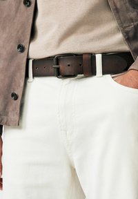 Brown leather belt with rectangular metal buckle, worn with light-colored pants and tan top, complemented by a gray button-up jacket.