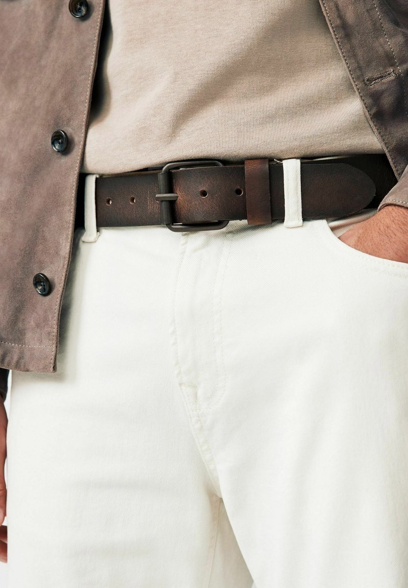 Brown leather belt with rectangular metal buckle, worn with light-colored pants and tan top, complemented by a gray button-up jacket.