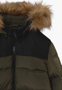 Olive green puffer jacket with black upper panels. Features a detachable brown fur hood and subtle logo detailing on the front.