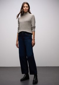 Femme debout portant un pull à col roulé beige clair, un jean large foncé et des bottines noires, sur un fond gris uni.