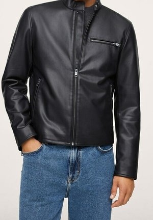 Homme portant une veste en cuir noire zippée avec une poche poitrine zippée et un jean bleu, mains partiellement dans les poches.