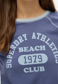 Sudadera azul marino con letras en azul claro. Presenta mangas raglán, cuello de canalé y texto impreso: "SUPERDRY ATHLETICS BEACH 1979 CLUB."