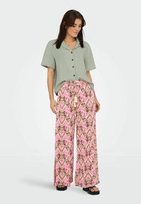 Femme portant une chemise à manches courtes boutonnée vert clair et un pantalon large à motifs roses avec des sandales noires, debout devant un fond uni.