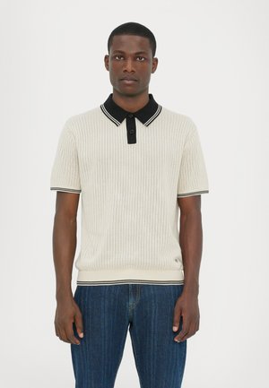 Cremefarvet ribbet polo shirt med en sort krave med hvide striber. Korte ærmer, knaplukning og tætsiddende stil. Båret sammen med blå jeans.