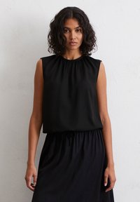 Vrouw met krullend donker haar, gekleed in een mouwloos zwart topje en een bijpassende rok, staande tegen een effen lichtgrijze muur.