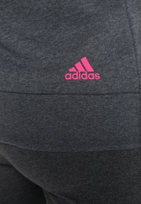 Grijze sportbroek met een gladde textuur en een roze Adidas-logo aan de zijkant. Het ontwerp heeft een aansluitende snit en een elastische tailleband.