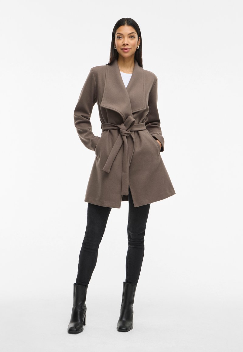 Abrigo marrón con un amplio cuello y cinturón, cuenta con bolsillos laterales y se combina con jeans skinny negros y botas de tobillo negras.