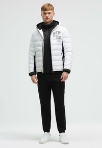 Chaqueta blanca acolchada con capucha negra y acentos laterales, con un estampado gráfico. Combinada con una sudadera negra con cremallera y pantalones negros.