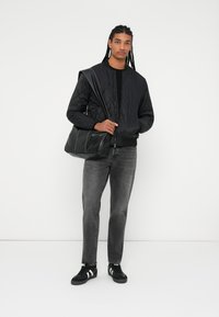 Quiltet sort bomberjakke, sort sweater, grå slim-fit jeans, sort skuldertaske. Modellen har sorte sneakers med hvide striber på.