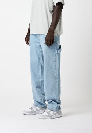 Vaqueros de denim azul claro con un corte holgado y detalles de bolsillos laterales, combinados con zapatillas grises y blancas. Camiseta gris lisa encima.