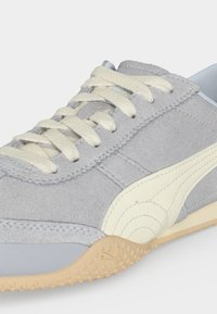 Γκρι σουέντ αθλητικό παπούτσι με κρεμ κορδόνια, κρεμ ρίγα Puma και υφασμένο μπεζ καουτσούκ στη σόλα, εμφανίζεται από κοντινή πλευρική γωνία.