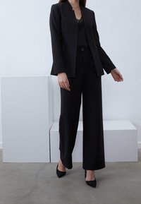 Zwarte op maat gemaakte blazer met een diepe V-hals, gedragen over een kanten top, gecombineerd met hoge taille zwarte broeken en puntige zwarte hakken.