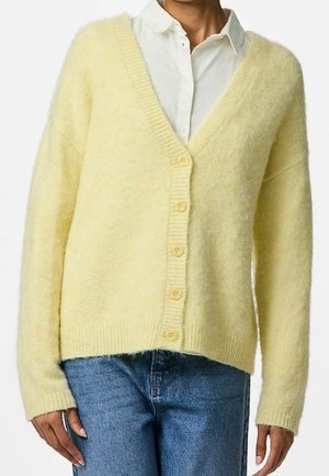 Osoba nosící světle žlutý měkký cardigan na knoflíky přes bílou košili s límečkem a modré džíny, zobrazená od krku po boky.