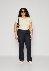 Top beige com riscas e mangas curtas, jeans flare de lavagem escura e sapatos brancos de salto com Acabamentos metálicos. Modelo a usar óculos de sol e uma mala preta.