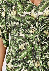 Groene tropische shirt met een patroon van grote palmbladeren en ananassen, met een twistdetail aan de voorkant en korte mouwen.