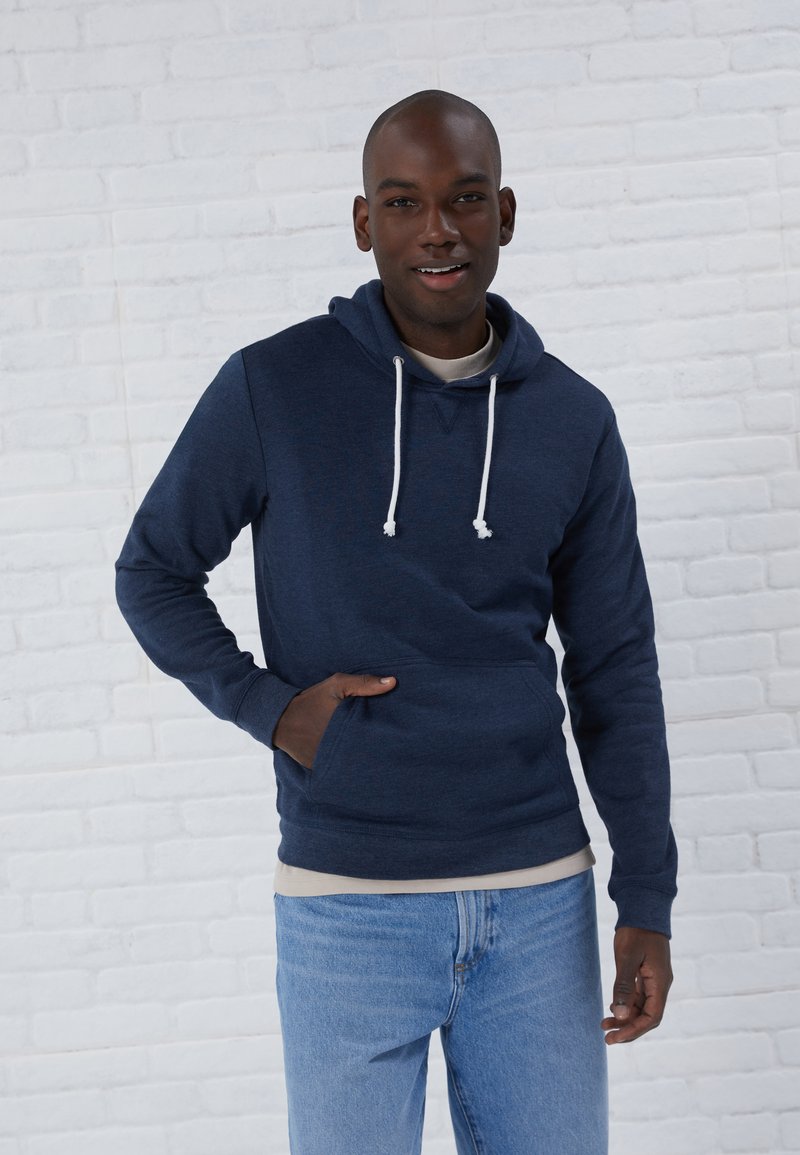 Marineblauwe hooded sweatshirt met een voorzak, witte trekkoorden en een beige binnenlaag, gedragen met lichtblauwe jeans tegen een achtergrond van witte bakstenen.