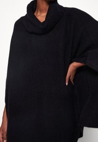 Czarny dzianinowy poncz z kominem, charakteryzujący się luźnym krojem i bocznymi kieszeniami. Tekstura wydaje się miękka i ciepła, idealna na chłodniejsze dni.