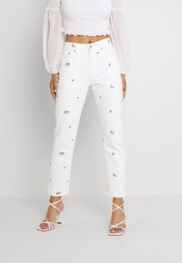 Weiße bestickte Jeans mit floralen Mustern, hochgeschnitten und mit geradem Bein, kombiniert mit einem gekürzten, strukturierten Oberteil und Riemchenschuhen.