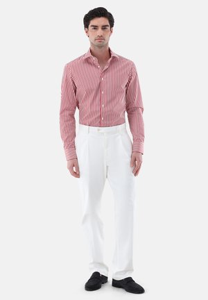 Mann trägt ein rot-weiß vertikal gestreiftes Langarmhemd, weiße Hose und schwarze Slipper, steht vor weißem Hintergrund.