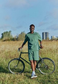 Haut vert surdimensionné à manches courtes et short assorti portés par une personne, debout à côté d'un vélo noir dans un champ d'herbe.