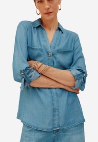 Camicia in denim azzurro chiaro con maniche lunghe, chiusura a bottoni frontale e due tasche sul petto; il tessuto ha una texture liscia e una vestibilità casual.